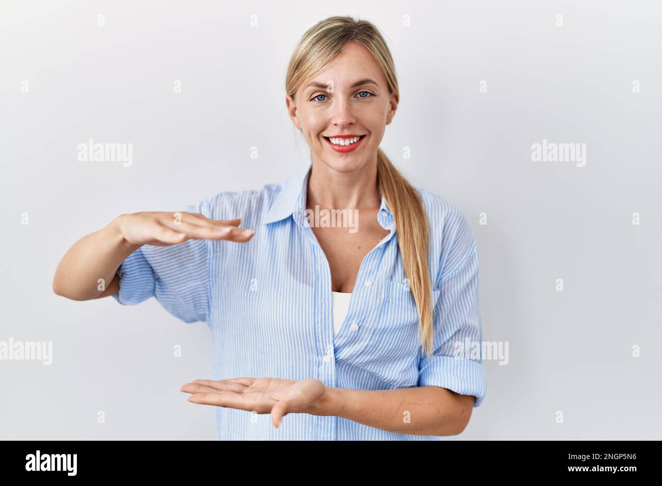 Hermosa mujer rubia de pie sobre fondo blanco gesticulando con las manos  que muestran signo grande y grande del tamaño, símbolo de la medida.  sonriendo mirando a la cam Fotografía de stock -