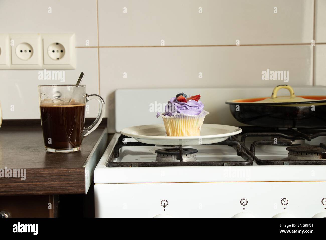 Una magdalena se coloca en una estufa de gas y al lado hay una taza de café en la cocina para el desayuno en casa, postre y café Foto de stock