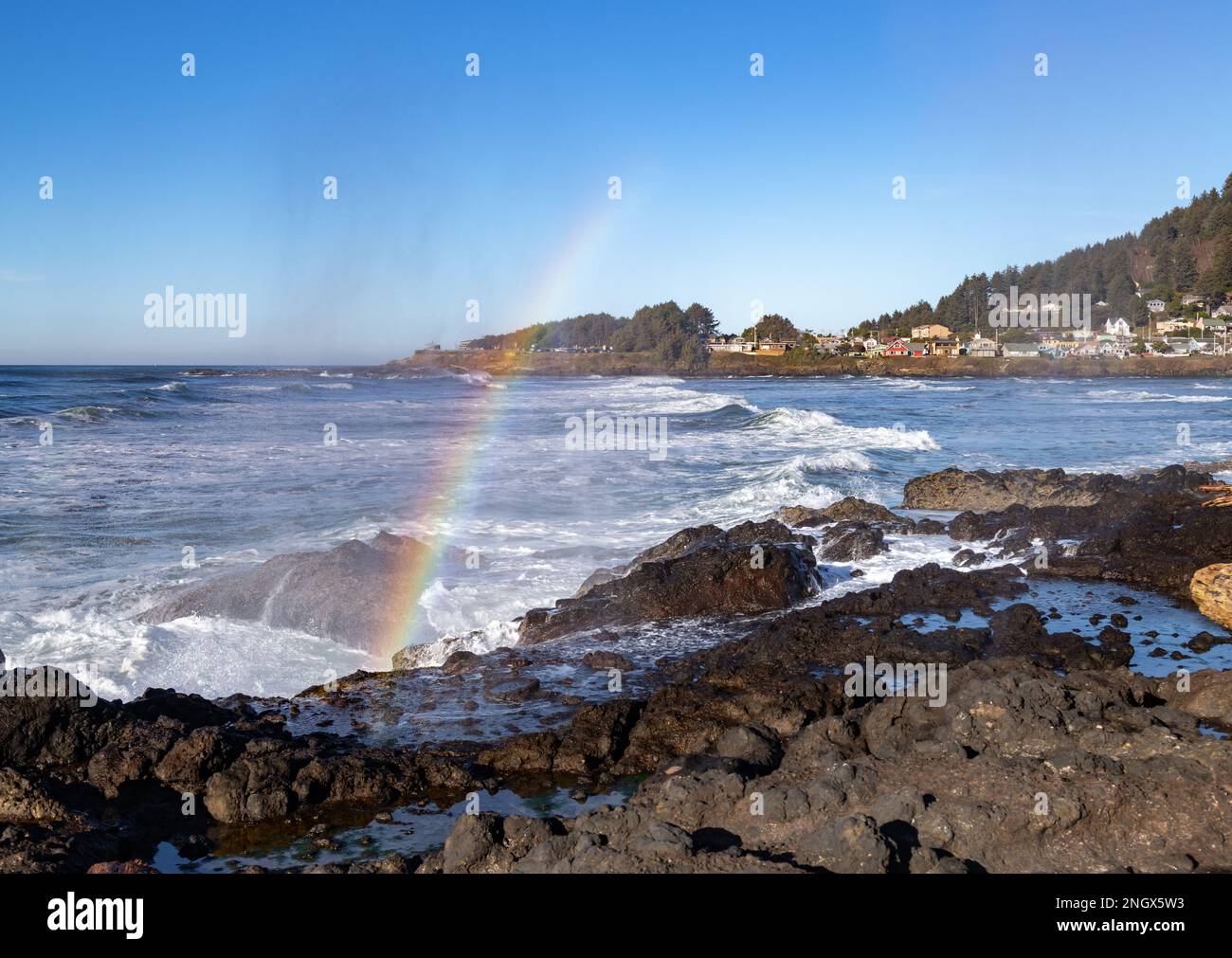 La ciudad de Oregon de Yachats en el fondo, con un arco iris en el primer plano, formado por la refracción de la luz del sol en la niebla de un cuerno de chorro Foto de stock