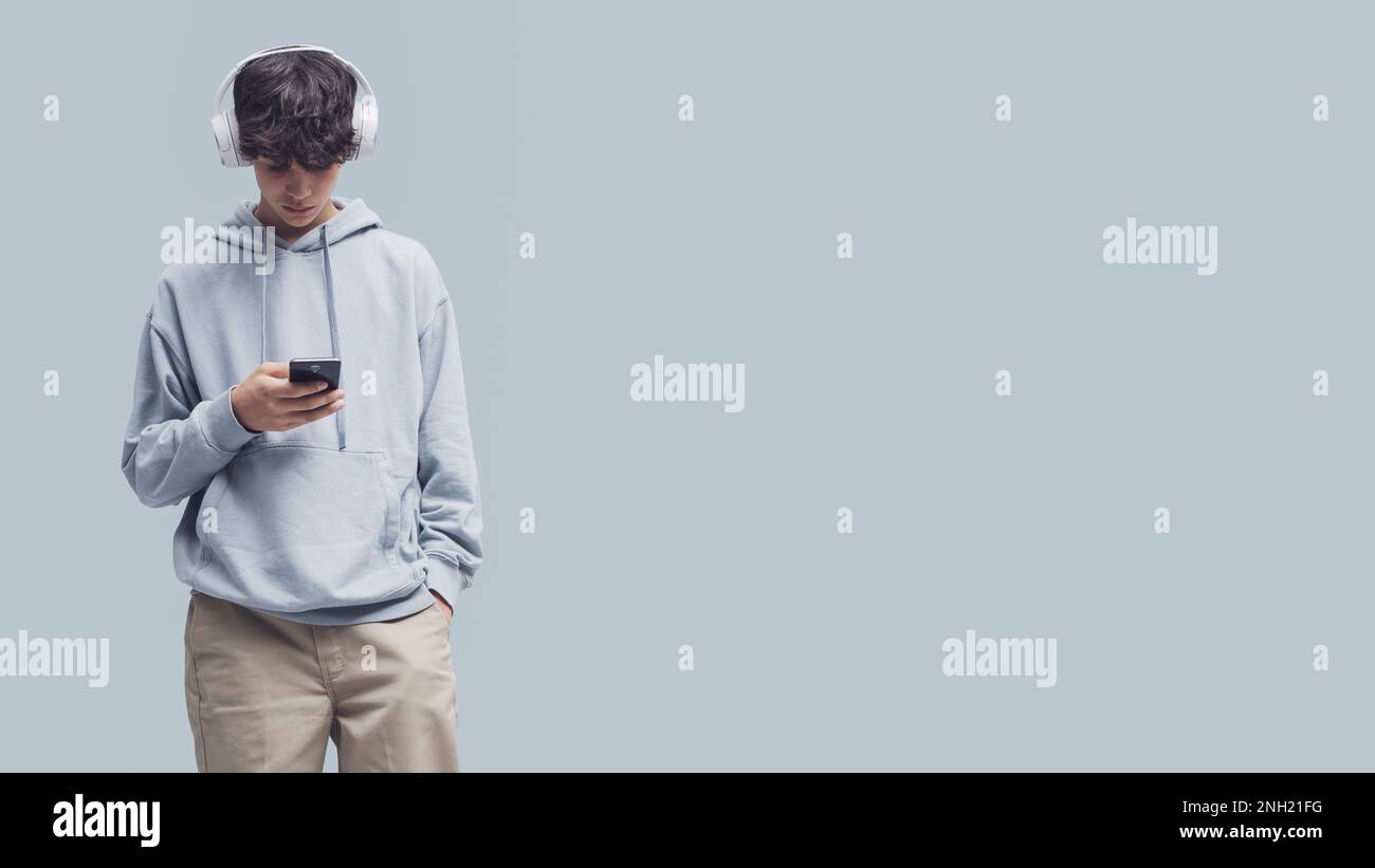 Adolescente fresco con auriculares y conexión con su teléfono inteligente, aislado en fondo gris, espacio de copia Foto de stock