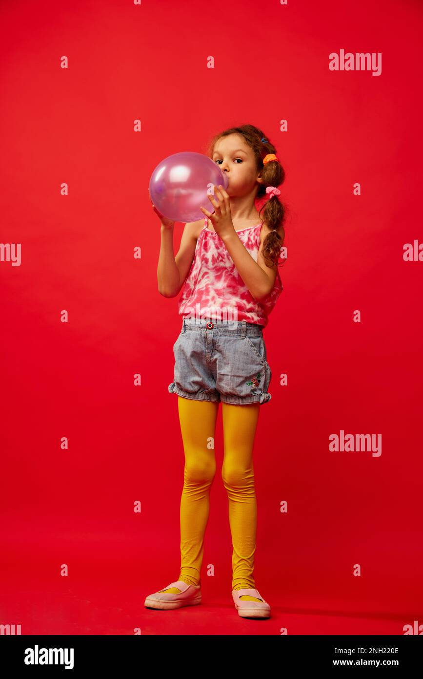 Pequeña niña linda, niño con trenzas posando emocionalmente en medias  amarillas, globo de aire soplado sobre fondo de estudio rojo brillante.  Concepto de infancia Fotografía de stock - Alamy