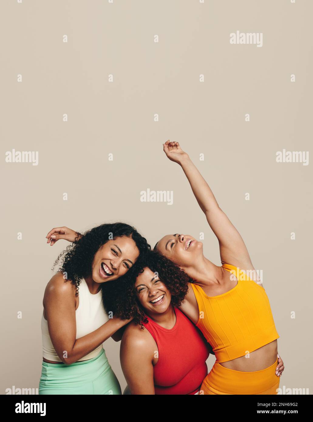 Tres mujeres jóvenes riendo alegremente mientras se paran juntas en un estudio con ropa deportiva. Grupo de amigas celebrando su forma, saludable Foto de stock