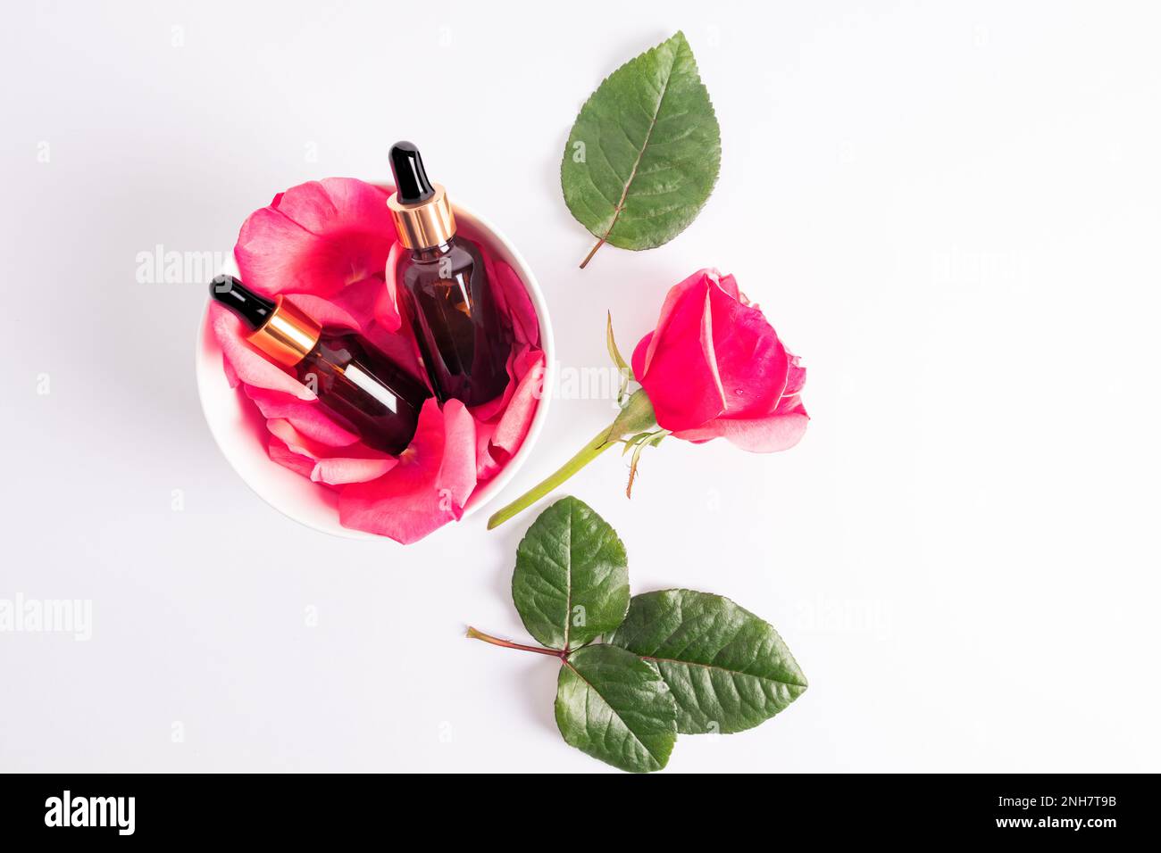 Dos botellas cosméticas con una tapa de oro y una pipeta con aceite de rosa natural o suero hidratante para el cuidado personal se encuentran en un tazón con pétalos de rosa. Vista superior Foto de stock