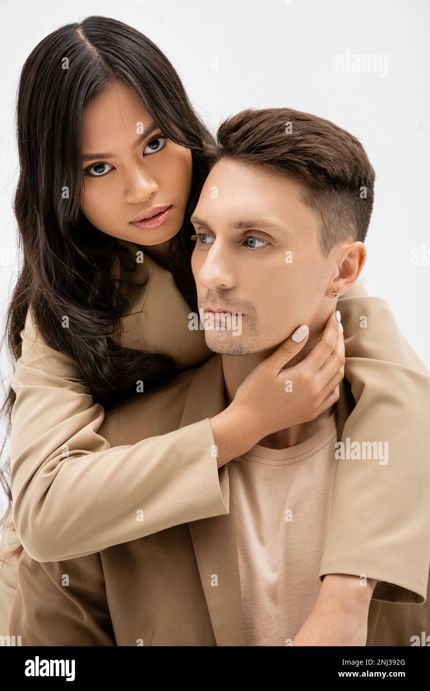 mujer asiática morena mirando a la cámara y abrazando al hombre en blazer beige aislado en gris, imagen de stock Fotografía de stock