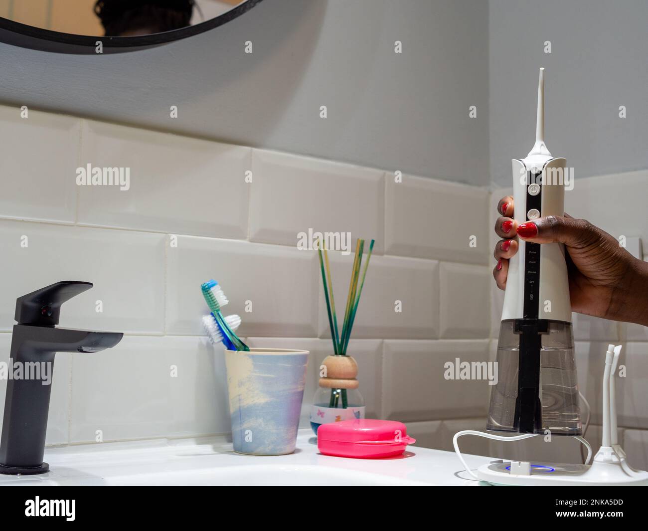 mujer joven que sostiene un hilo dental inalámbrico doméstico del agua en el lavabo del cuarto de baño Foto de stock