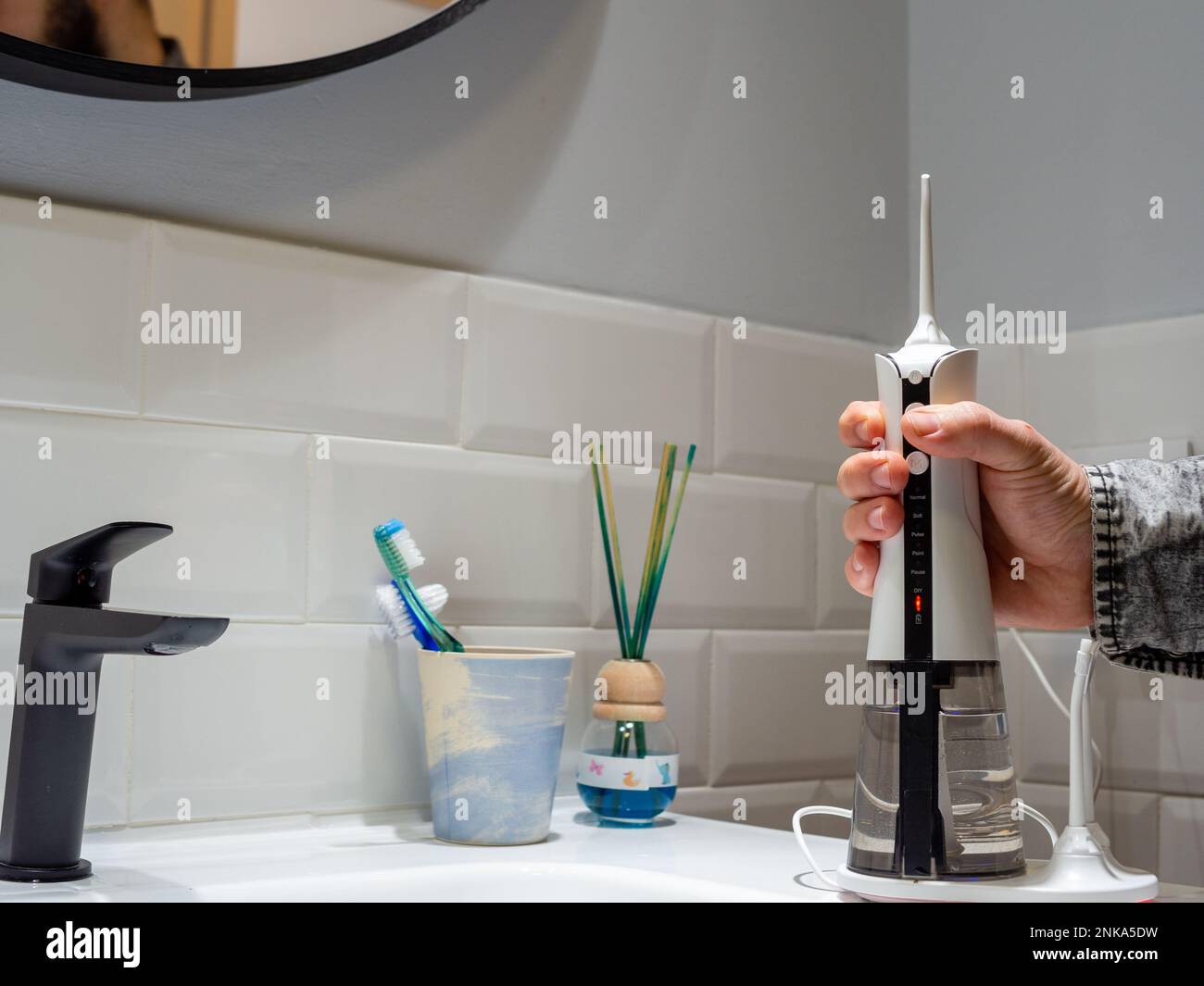 hombre joven que sostiene un hilo dental inalámbrico doméstico del agua en el lavabo del cuarto de baño Foto de stock