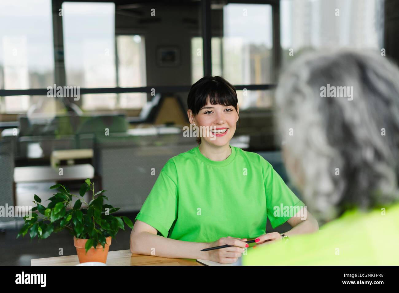 Mujer sonriente en camiseta verde mirando a colega en la oficina Foto de stock