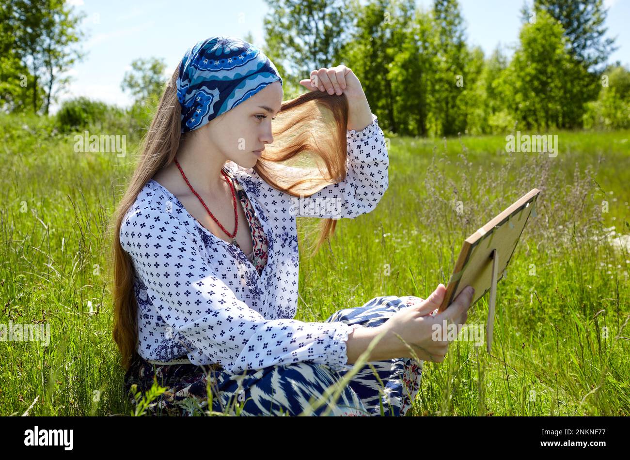 Mujer sentada en la hierba mirando su imagen del espejo. Retrato de chica atractiva en vestido en un día soleado de verano Foto de stock