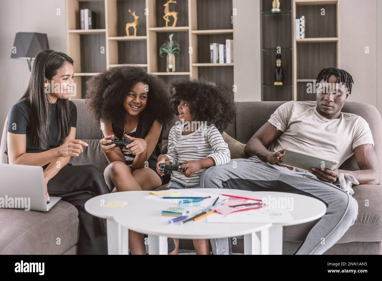 Familia africana negra feliz disfrutan de vacaciones juntos en el sofá sala de estar Dos hijas jugando al juego madre en línea trabajando con computadora portátil y baño de papá Foto de stock