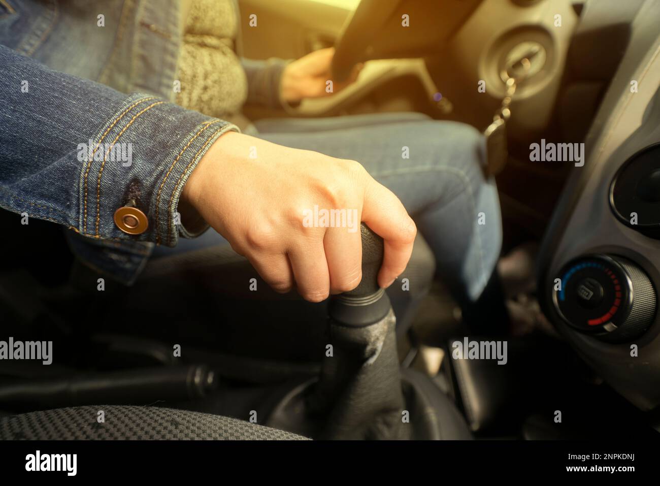 Cerca de la mano de una mujer vestida de azul en la palanca de cambios mientras conduce dentro de su vehículo iluminado con luz amarilla Foto de stock