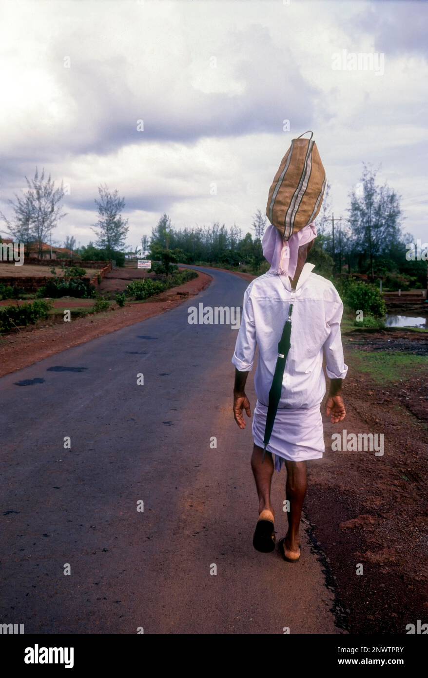 Un hombre que lleva una bolsa sobre su cabeza y un paraguas detrás de su camisa en la carretera, Kerala, India, Asia Foto de stock