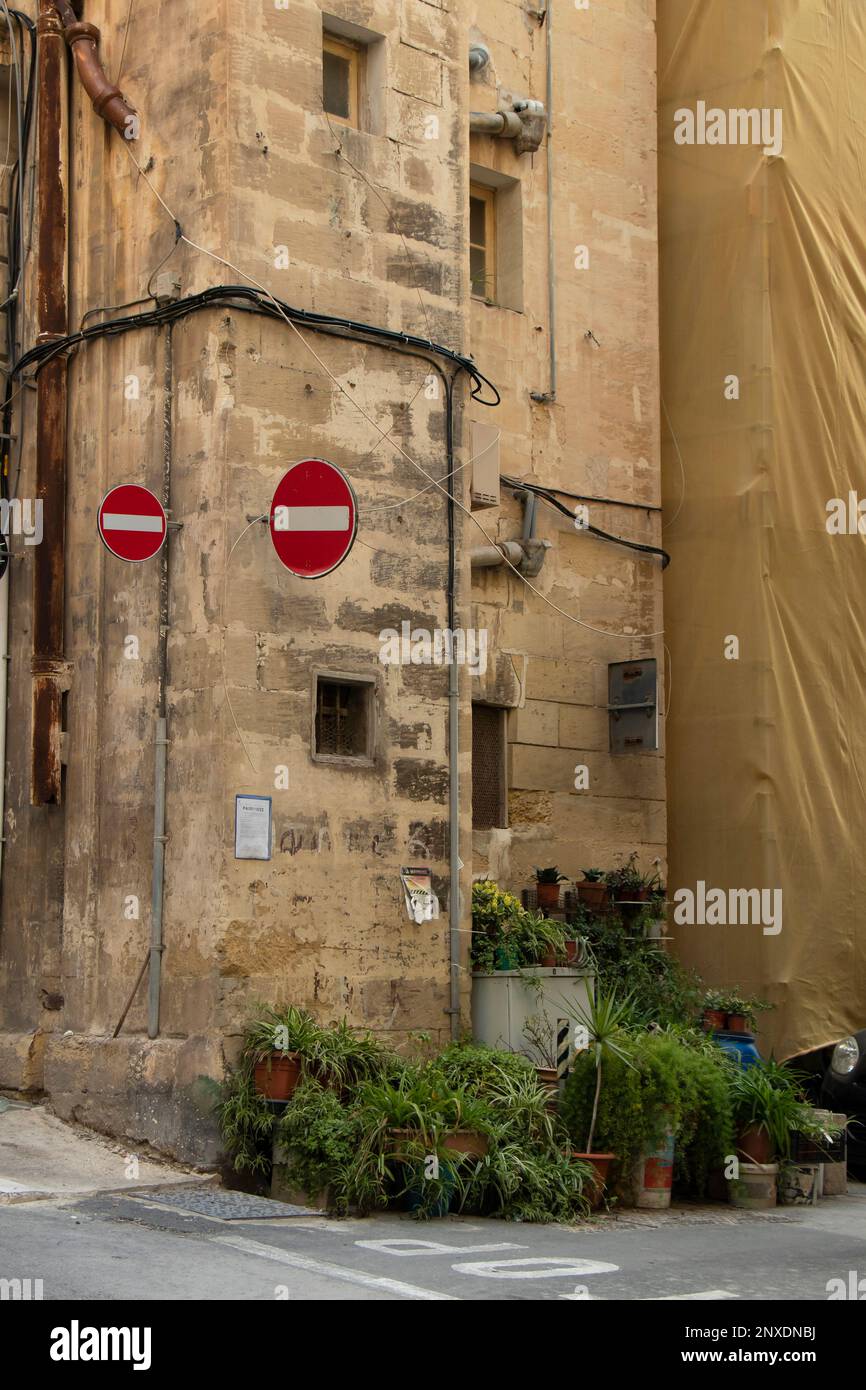 Valletta, Malta - 11 de noviembre de 2022: Casa de esquina de piedra caliza con dos señales de tráfico prohibitivas que marcan la dirección equivocada y plantas de maceta en la acera Foto de stock