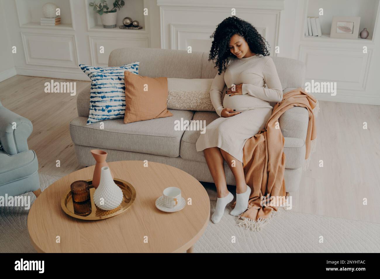 Hermosa foto de mujer afro americana embarazada sentada en un sofá acogedor decorado con almohadas de colores colocando suavemente las manos en su estómago mientras se relaja Foto de stock