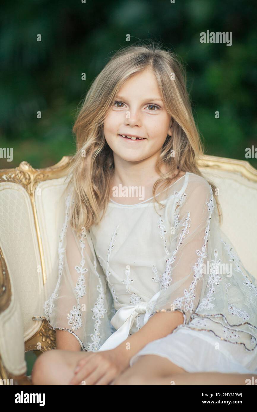 Pequeña chica rubia descalza en vestido blanco lujo sentado en crema y silla antigua oro en jardín Fotografía stock -
