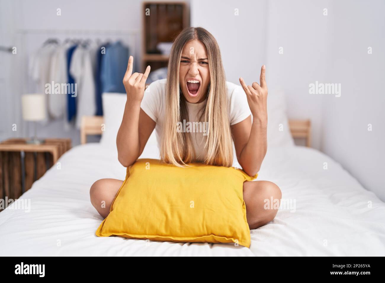 Mujer rubia joven que se sienta en la cama con la almohada en casa que  grita con expresión loca que hace el símbolo de la roca con las manos para  arriba. estrella
