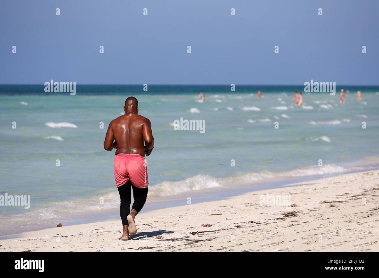 Hombre negro descalzo corriendo por la arena en el fondo de las olas del mar. Ejercicio en una playa, estilo de vida saludable Foto de stock