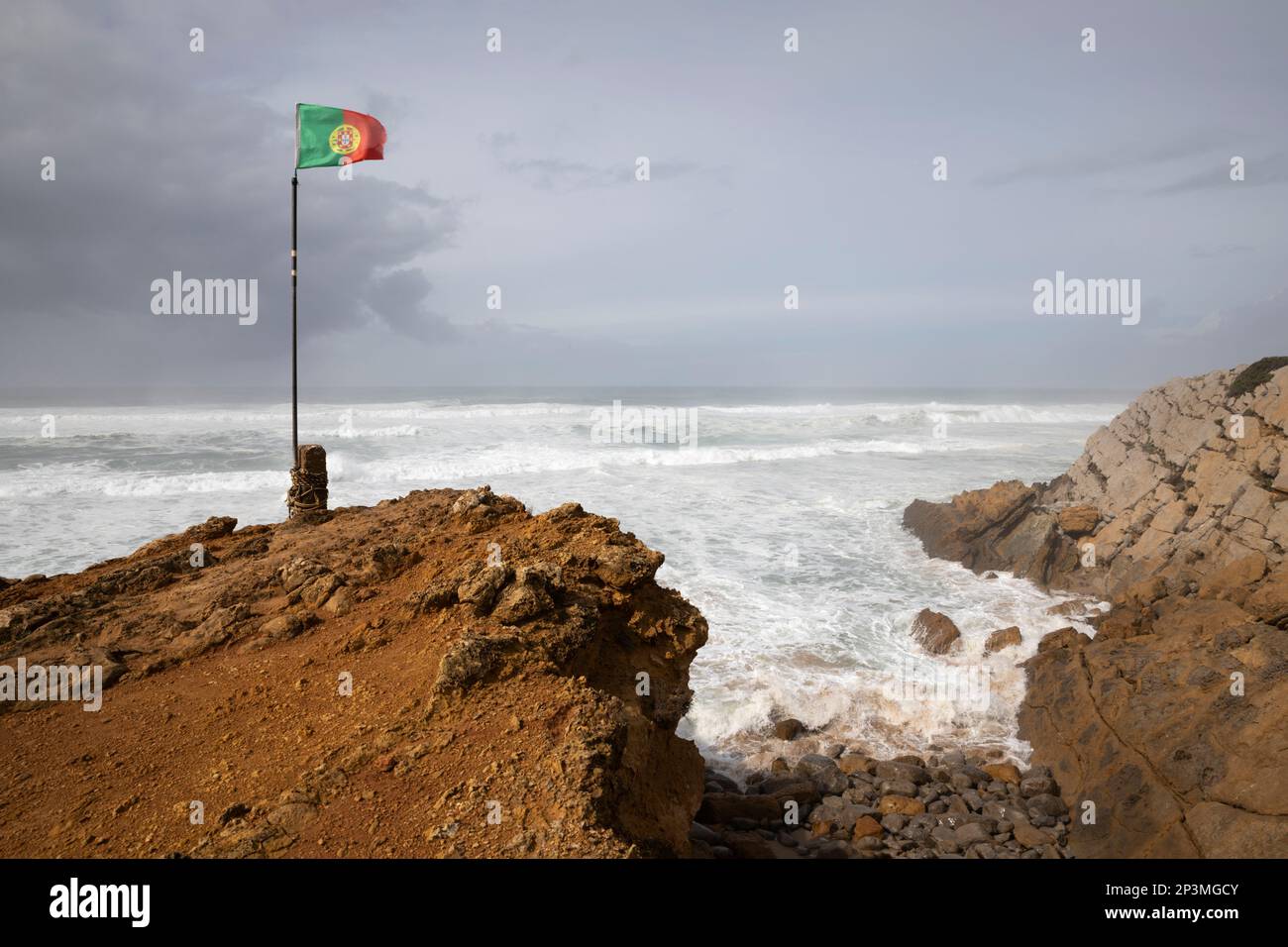 Bandera portuguesa volando en viento con las olas tormentosas del océano Atlántico rompiendo en la playa de Praia do Guincho Foto de stock