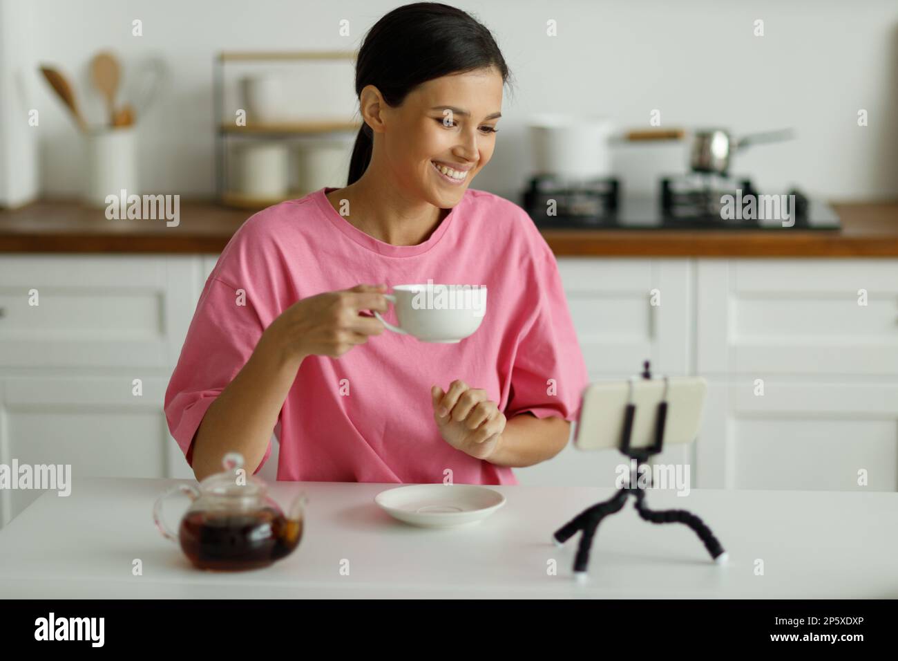 La mujer morena joven sonriente atractiva en la camiseta oversize rosa está bebiendo té en la cocina. Muchacha bonita feliz con la sonrisa blanca como la nieve saluda amigo, co Foto de stock