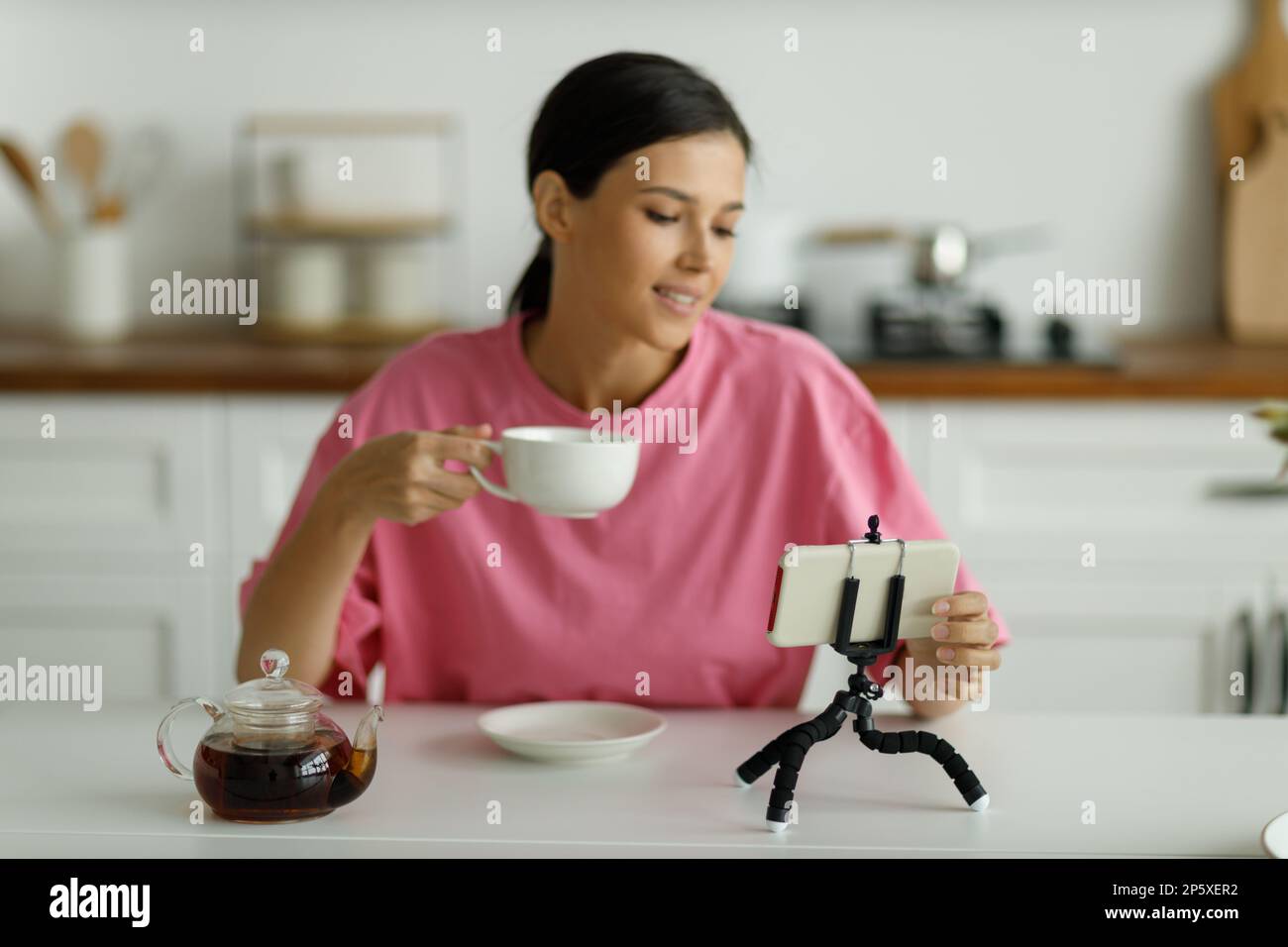 La mujer morena joven sonriente atractiva en la camiseta oversize rosa está bebiendo té en la cocina. Muchacha bonita feliz con la sonrisa blanca como la nieve saluda amigo, co Foto de stock