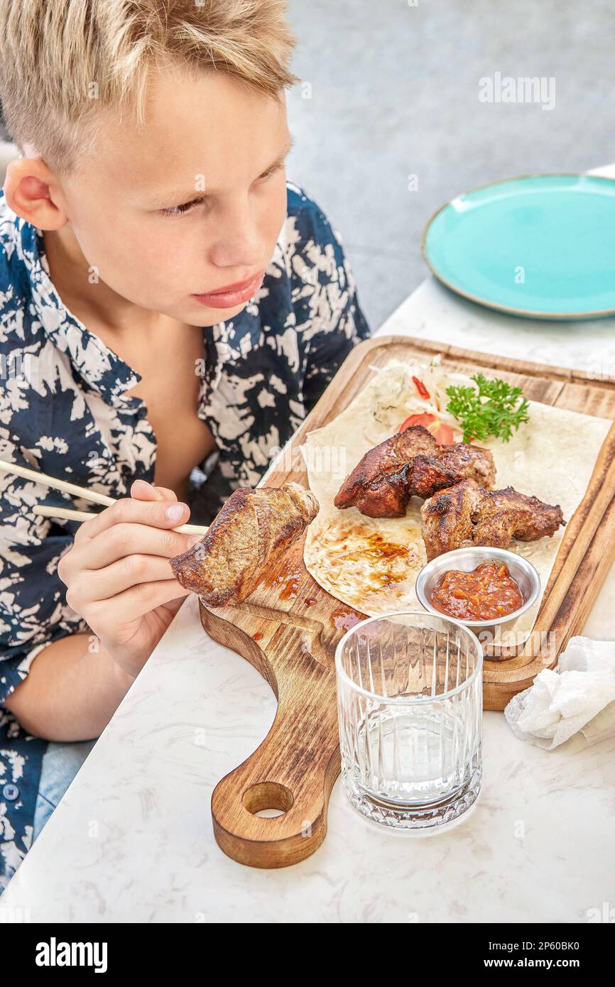 Chico rubio come plato de carne a la parrilla sentado en la mesa en el restaurante cerca de la vista superior. El niño preadolescente disfruta de una comida nutritiva en la cafetería Foto de stock