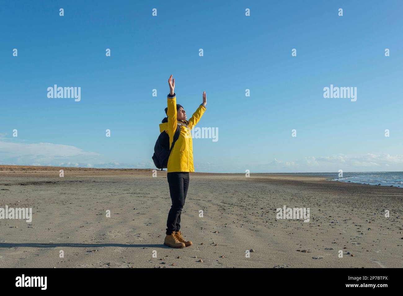 Mujer en un abrigo amarillo con los brazos levantados disfrutando del sol de invierno mientras camina en una playa Foto de stock