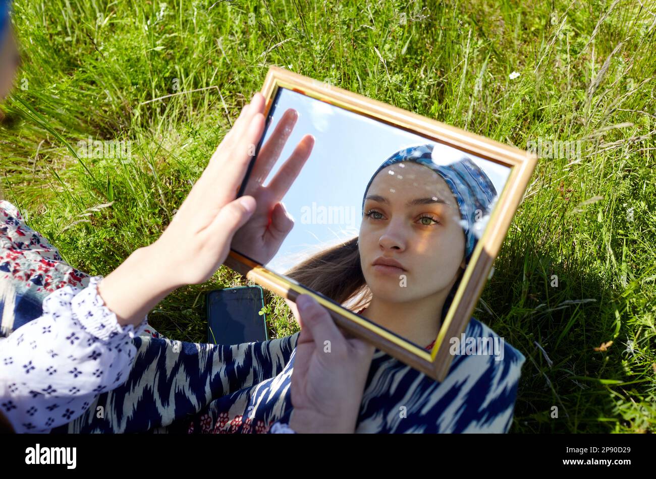 Mujer sentada en la hierba mirando su imagen del espejo. Retrato de chica atractiva en vestido en un día soleado de verano Foto de stock