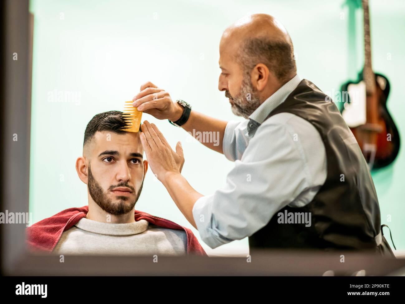 Vista en espejo de barbero barbudo concentrado peinando clientes cabello oscuro con peine amarillo Foto de stock