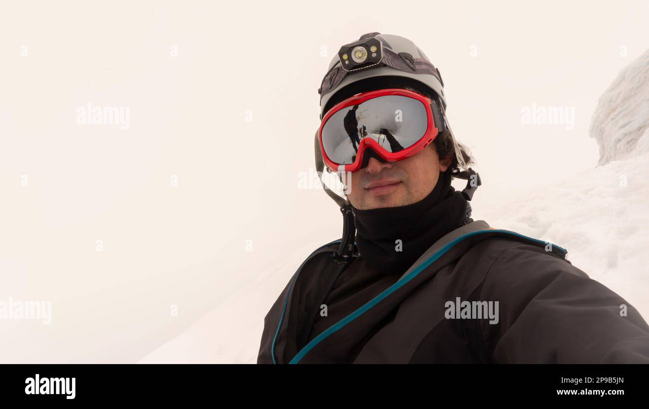 Escalador con casco, faro y chaqueta negra tomando un selfie en un glaciar en un día nublado, paisaje reflejado en gafas enmarcadas rojas Foto de stock