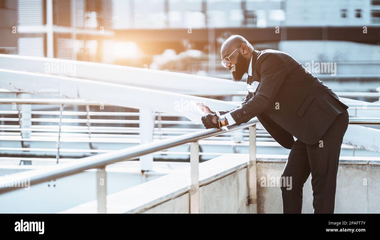 Vista de un hombre afro negro calvo maduro de lujo en un traje negro completo a medida, anteojos y corbata apoyados en la barandilla para una mejor lectura del papeleo t Foto de stock