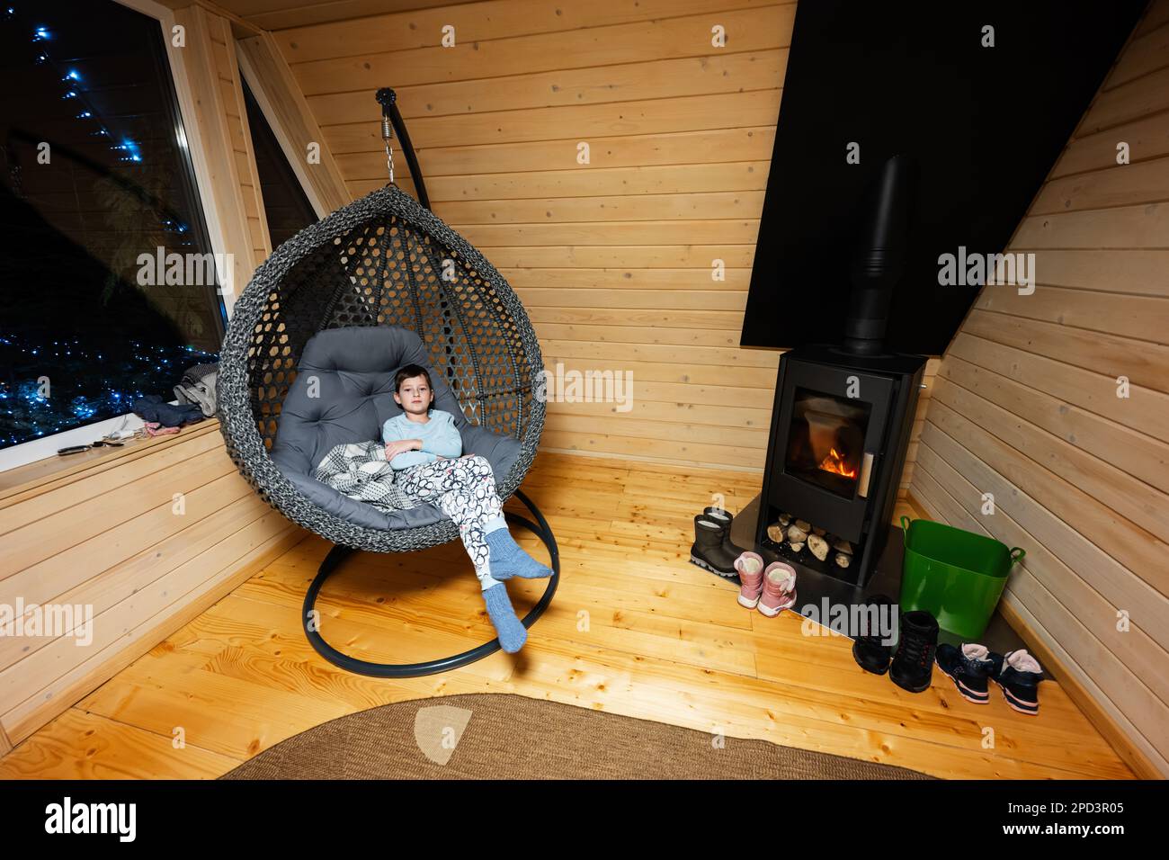 Niño en pijama y calcetines de punto en cómoda silla colgante contra la  chimenea en acogedora casita de madera pequeña cabaña. La vida en el campo  Fotografía de stock - Alamy