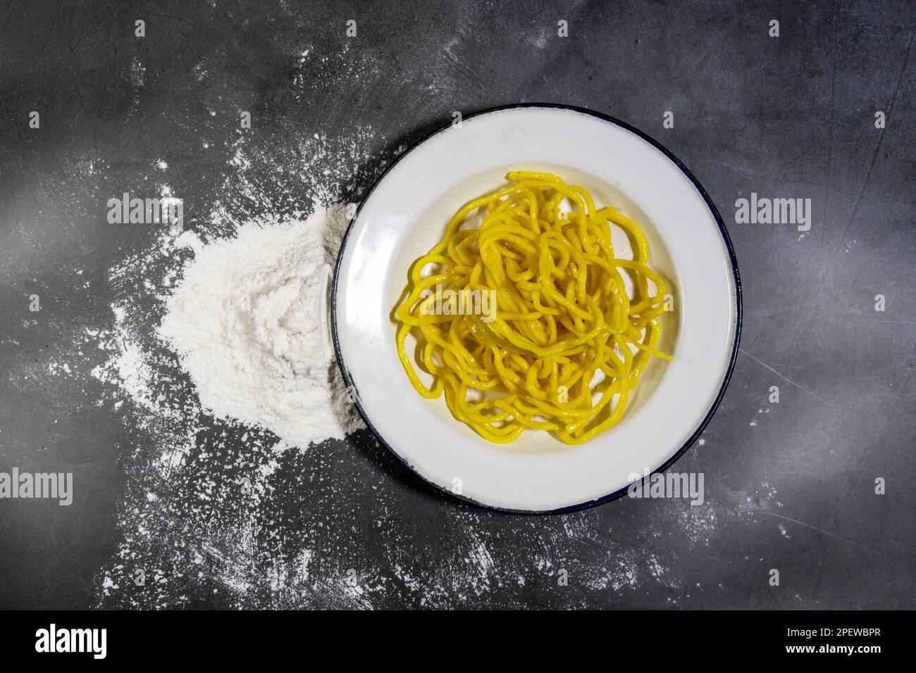 Vista superior de fideos amarillos en un plato blanco y harina de uso general, sobre el fondo oscuro. Foto de stock