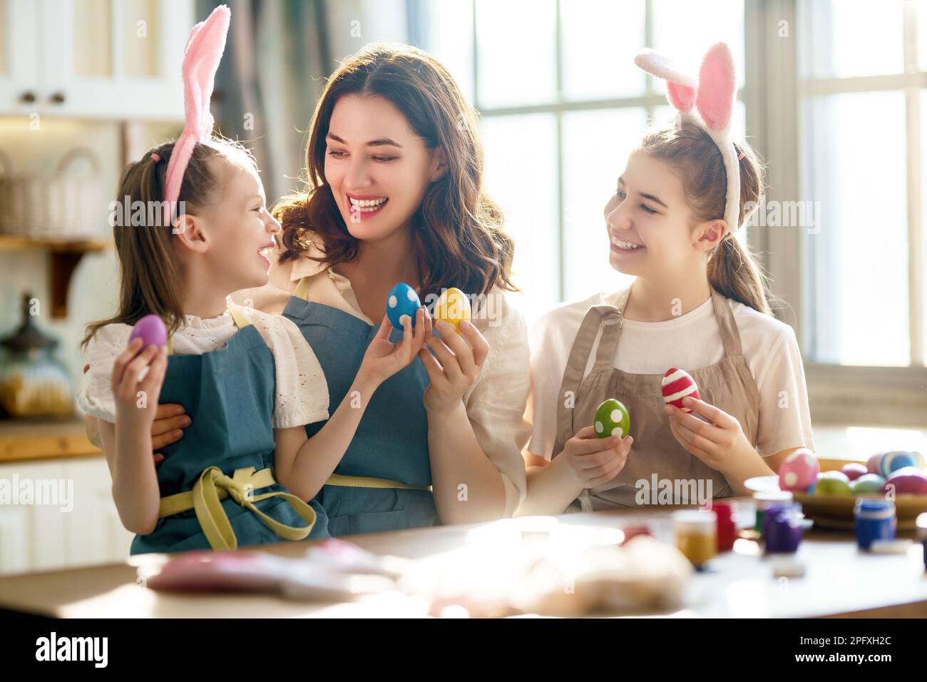 Madre feliz y sus hijas con delantales sosteniendo huevos pintados de  colores mientras los decoran con tintes de comida en la acogedora cocina en  casa. Artesanía de Pascua Fotografía de stock -
