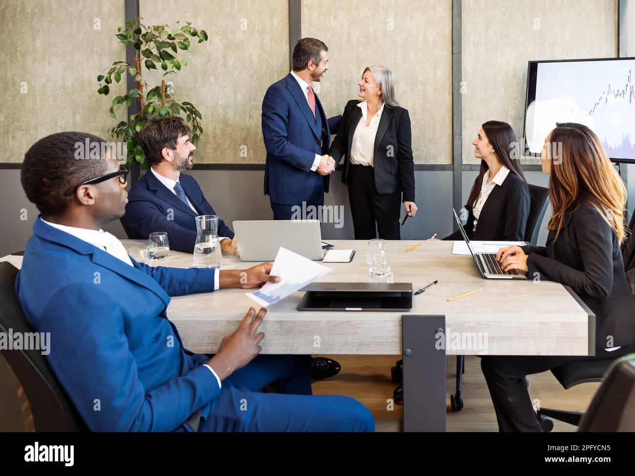 Grupo de empresarios diversos en ropa formal sentado en la mesa y mirando a los compañeros de trabajo estrechando la mano durante la reunión de negocios en la oficina moderna Foto de stock