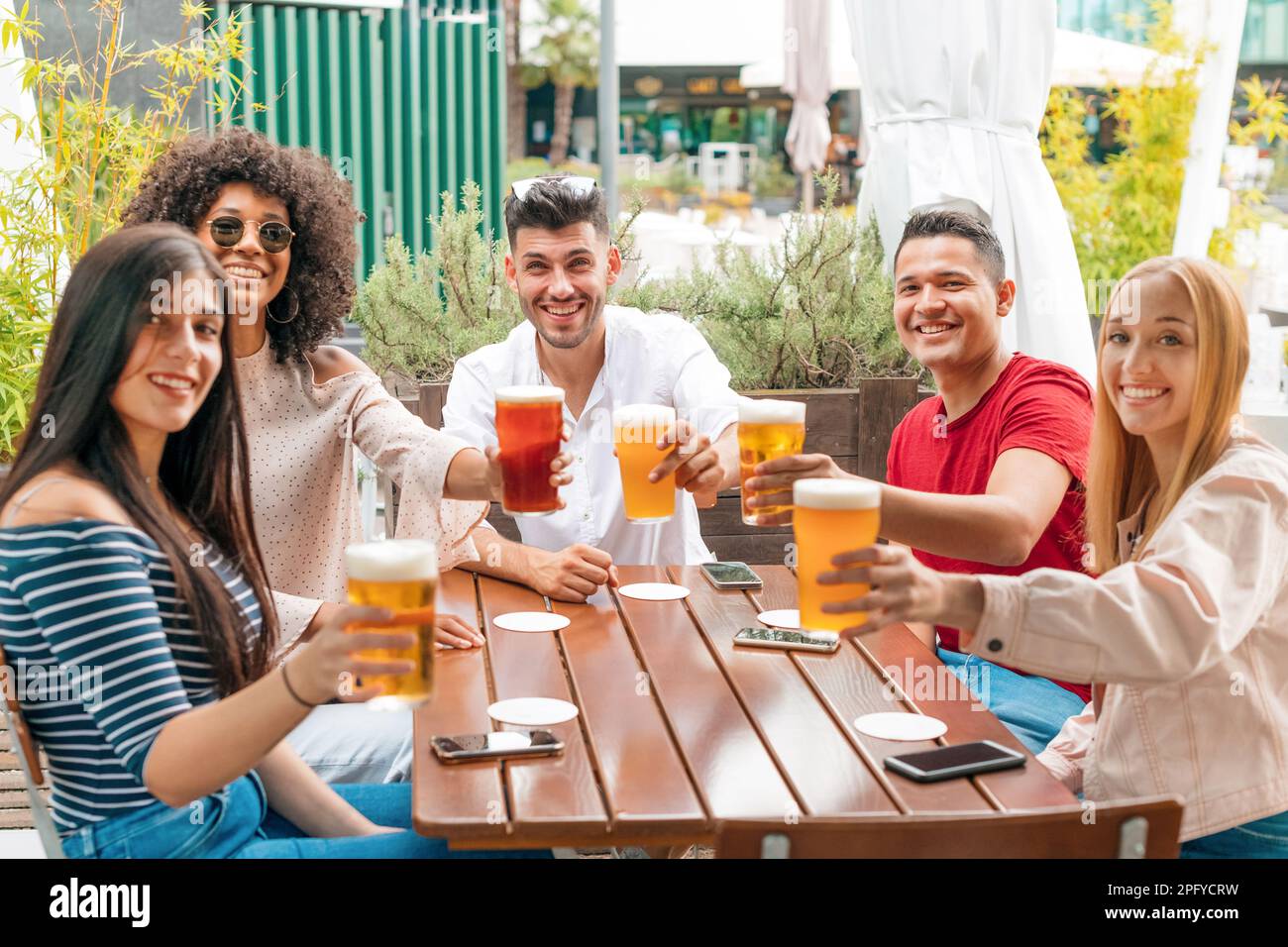 Grupo de amigos multiétnicos mirando hacia otro lado y animando con vasos de cerveza espumosos en la mesa de madera en el edificio de la terraza el día de verano Foto de stock