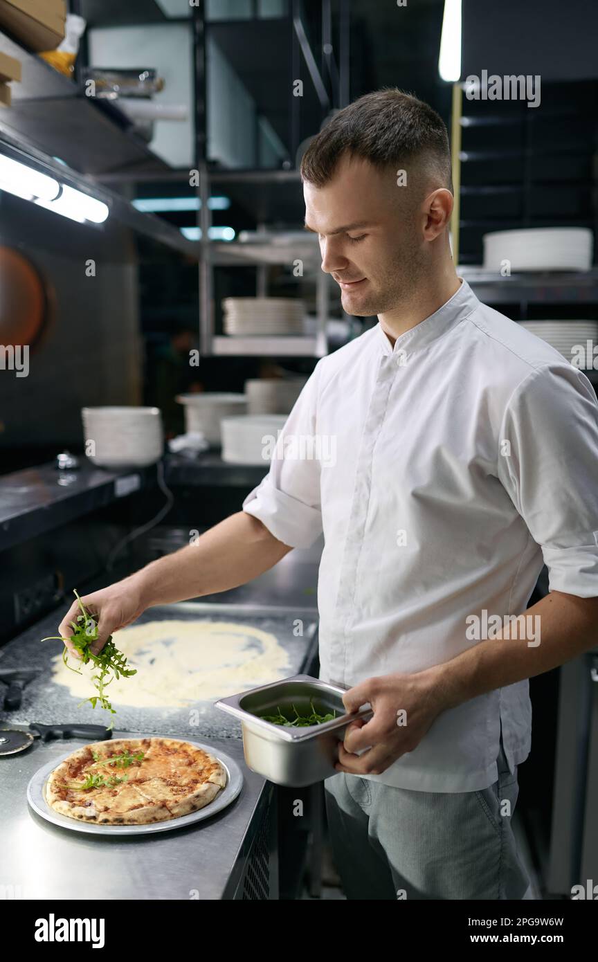 Chef masculino espolvorea vegetación fresca sobre pizza tradicional Foto de stock
