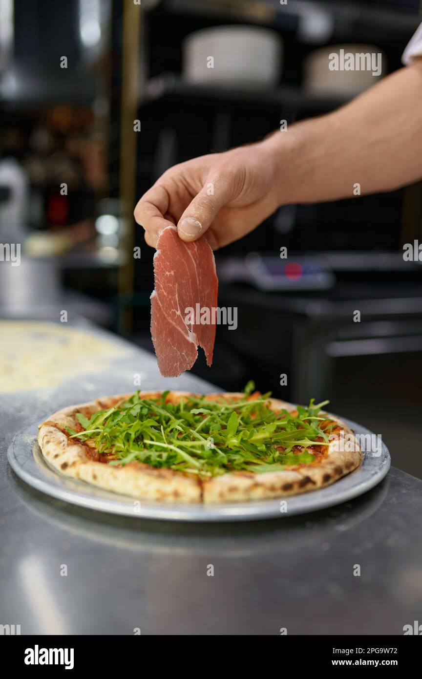 Primer plano de la mano del chef sosteniendo una rebanada de tocino sobre la pizza Foto de stock