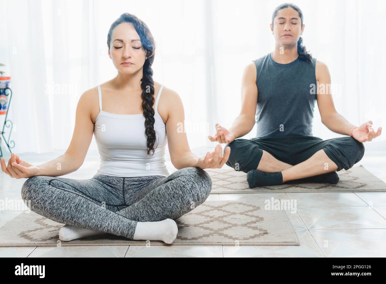 Dos personas de mediana edad están haciendo yoga en interiores, con mucha luz solar entrando por la ventana, haciendo posiciones de meditación profunda. Hay mucho de Foto de stock