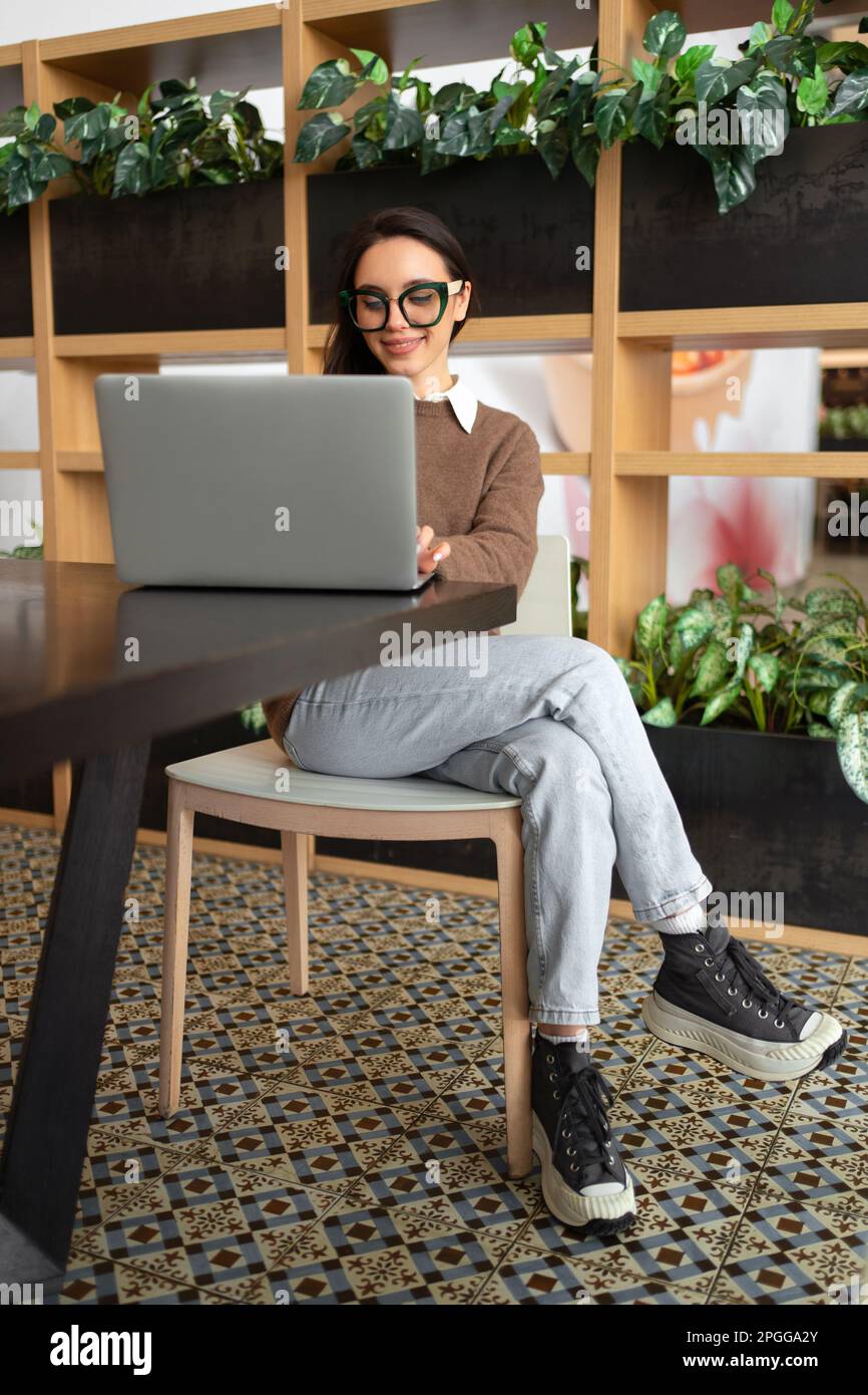 Mujer con gafas usando laptop. Empresaria encantadora en anteojos y ropa  casual que trabaja con la pantalla de observación del ordenador portátil.  Elegante mujer de negocios de longitud completa tiro vertical Fotografía