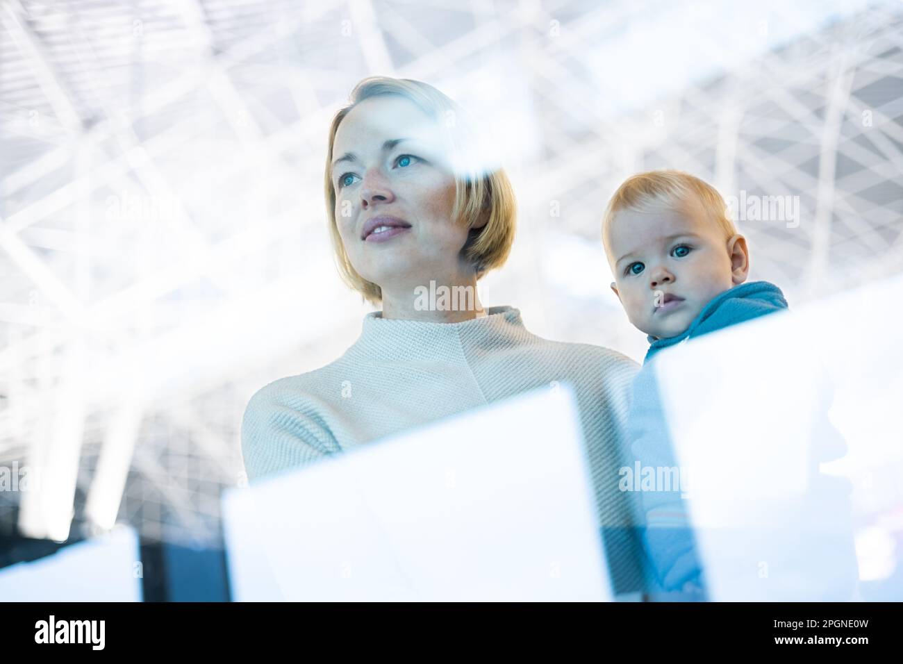 Madre joven pensativa que mira la ventana del abrevadero que sostiene a su niño bebé mientras espera para abordar un avión en las puertas de salida de la terminal del aeropuerto Foto de stock