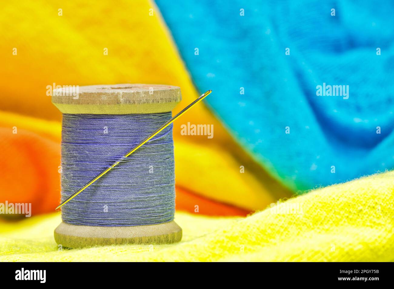 Carrete de hilo de algodón púrpura y aguja de coser sentado en ropa de color brillante. Concepto de sastrería textil con enfoque en primer plano. Foto de stock