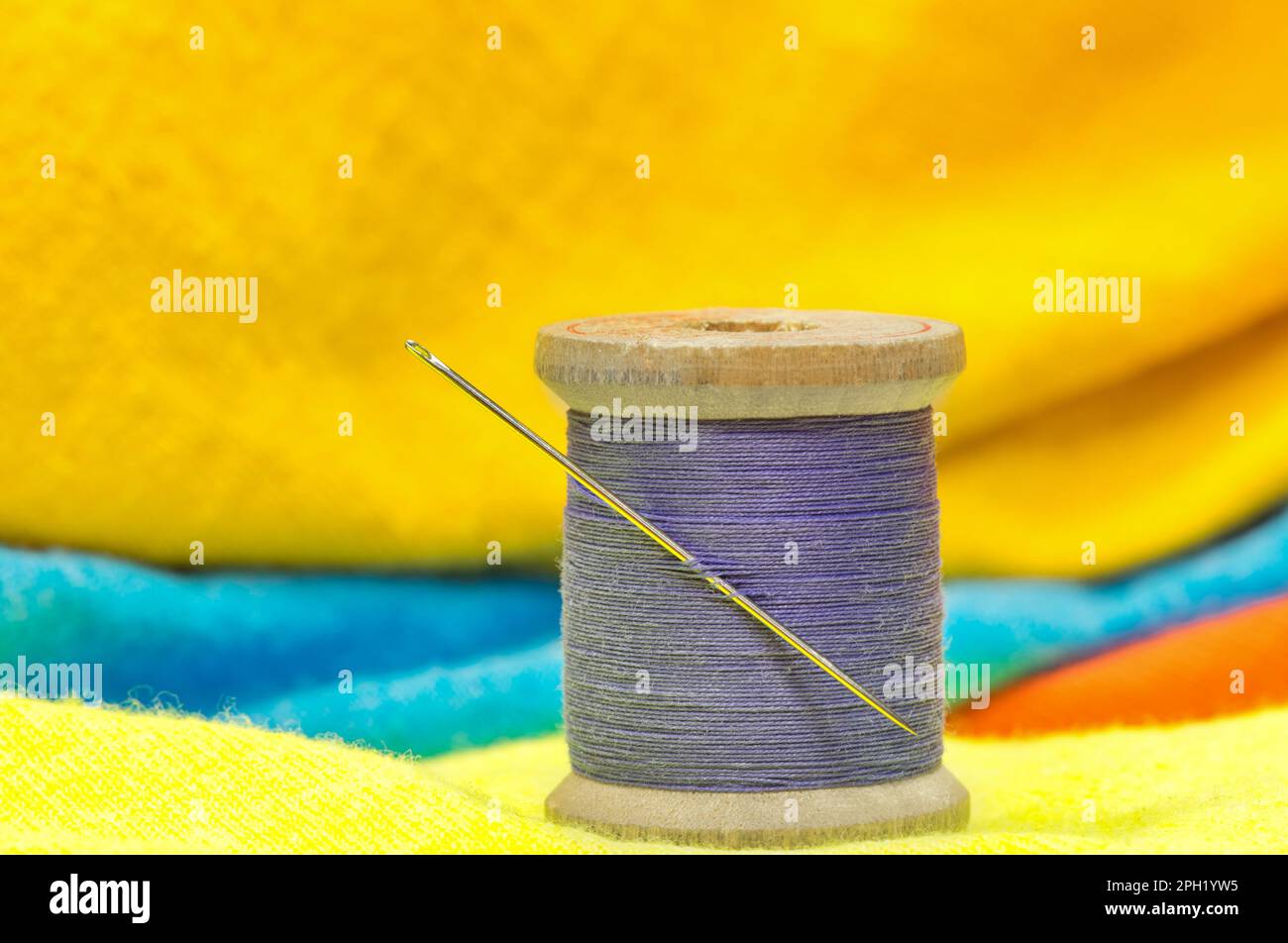 Carrete de hilo de algodón púrpura y aguja de coser sentado en ropa de color brillante. Concepto de sastrería textil con enfoque en primer plano. Foto de stock