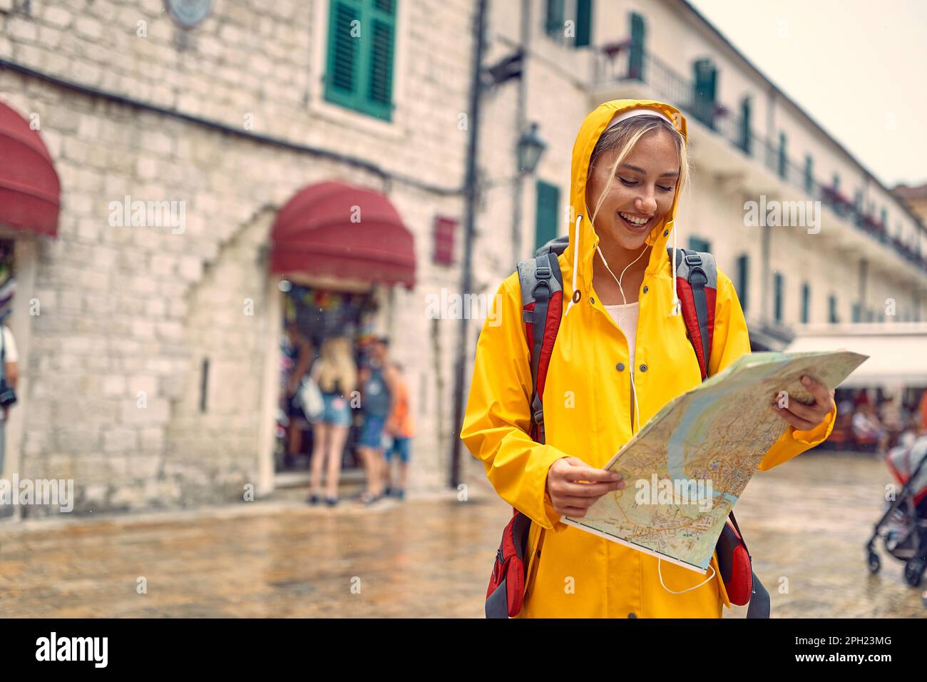 Mujer joven en chubasquero en la calle con el mapa, sintiéndose alegre. Lluvia de verano. Viaje, concepto de estilo de vida. Foto de stock