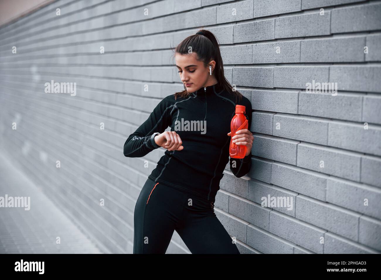 Chica deportiva joven en ropa deportiva negra de pie al aire libre cerca de  la pared gris con botella de agua y tomando un descanso Fotografía de stock  - Alamy