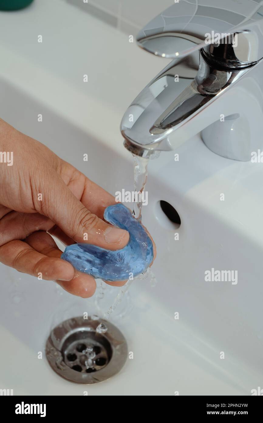 primer plano de un hombre enjuagando su férula oclusal azul bajo un chorro de agua de un grifo del lavabo del baño Foto de stock