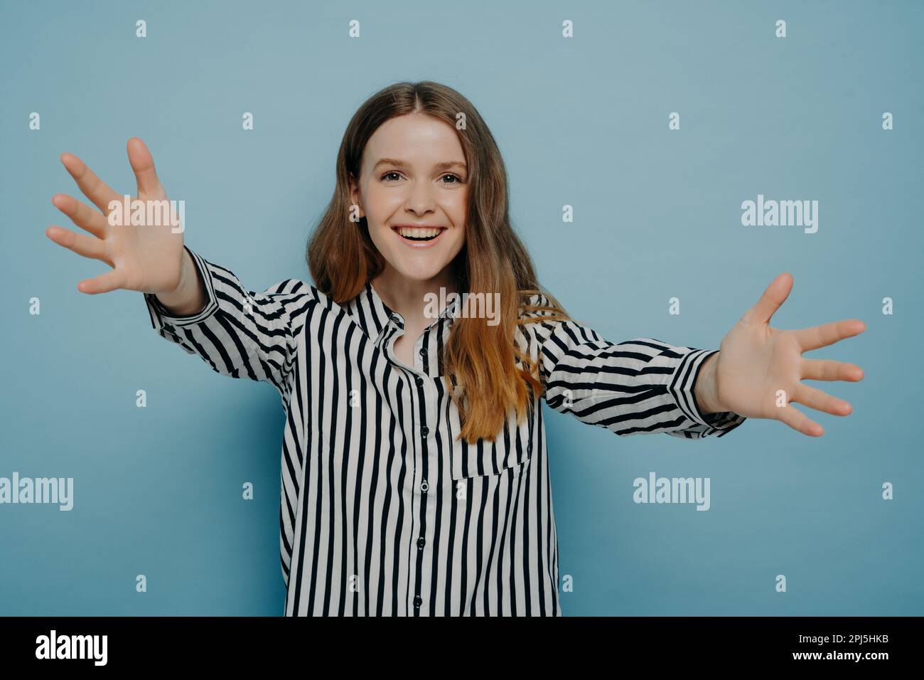 Retrato de mujer joven amorosa que ofrece un abrazo, sonriendo y tirando de las manos hacia la cámara mientras está de pie aislado sobre fondo azul, alegre adolescente gir Foto de stock