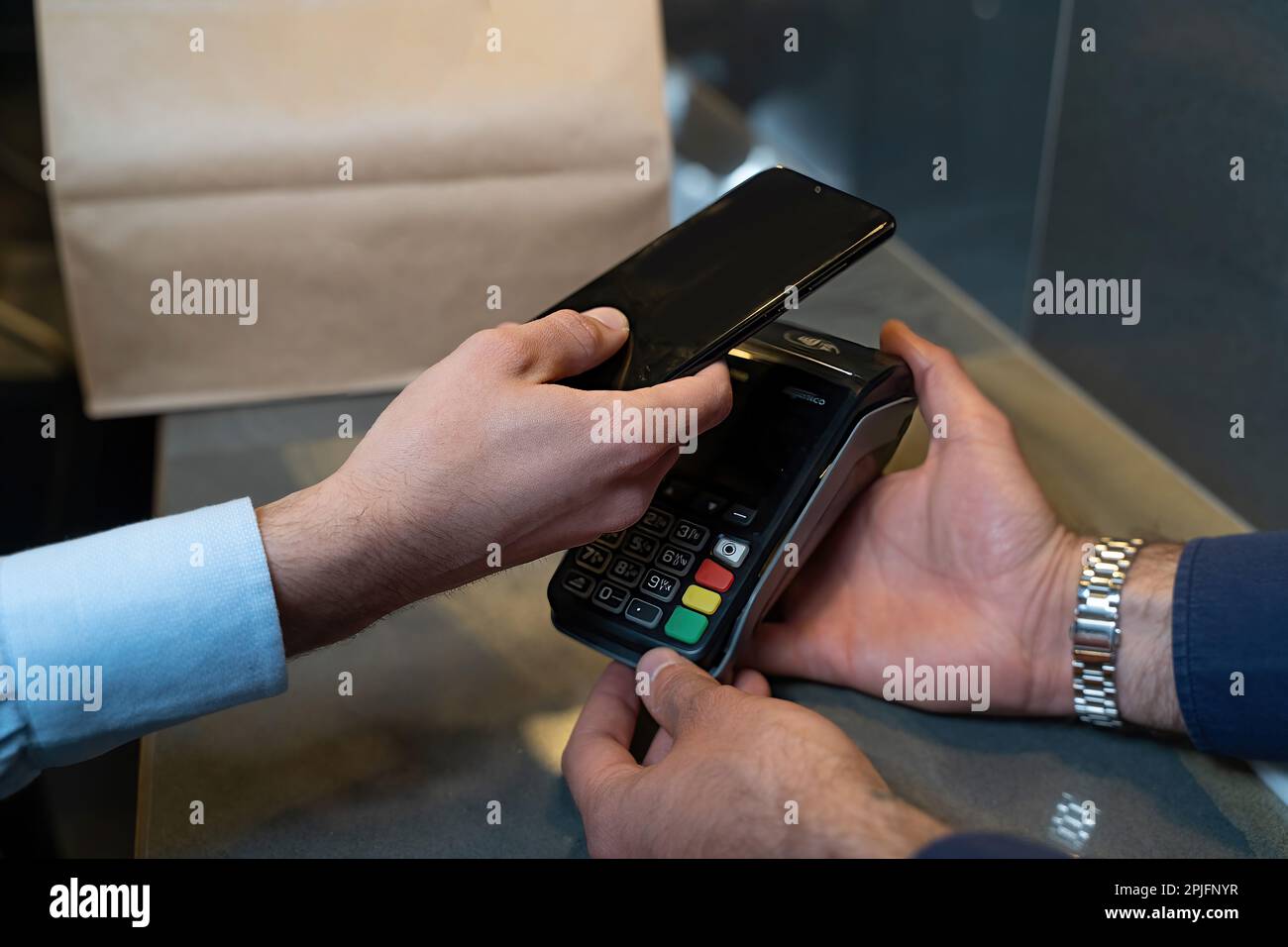 Un primer plano de las manos de dos personas, ya que una sostiene un lector  de tarjetas de crédito con un teclado, mientras que la otra utiliza un  teléfono inteligente para hacer
