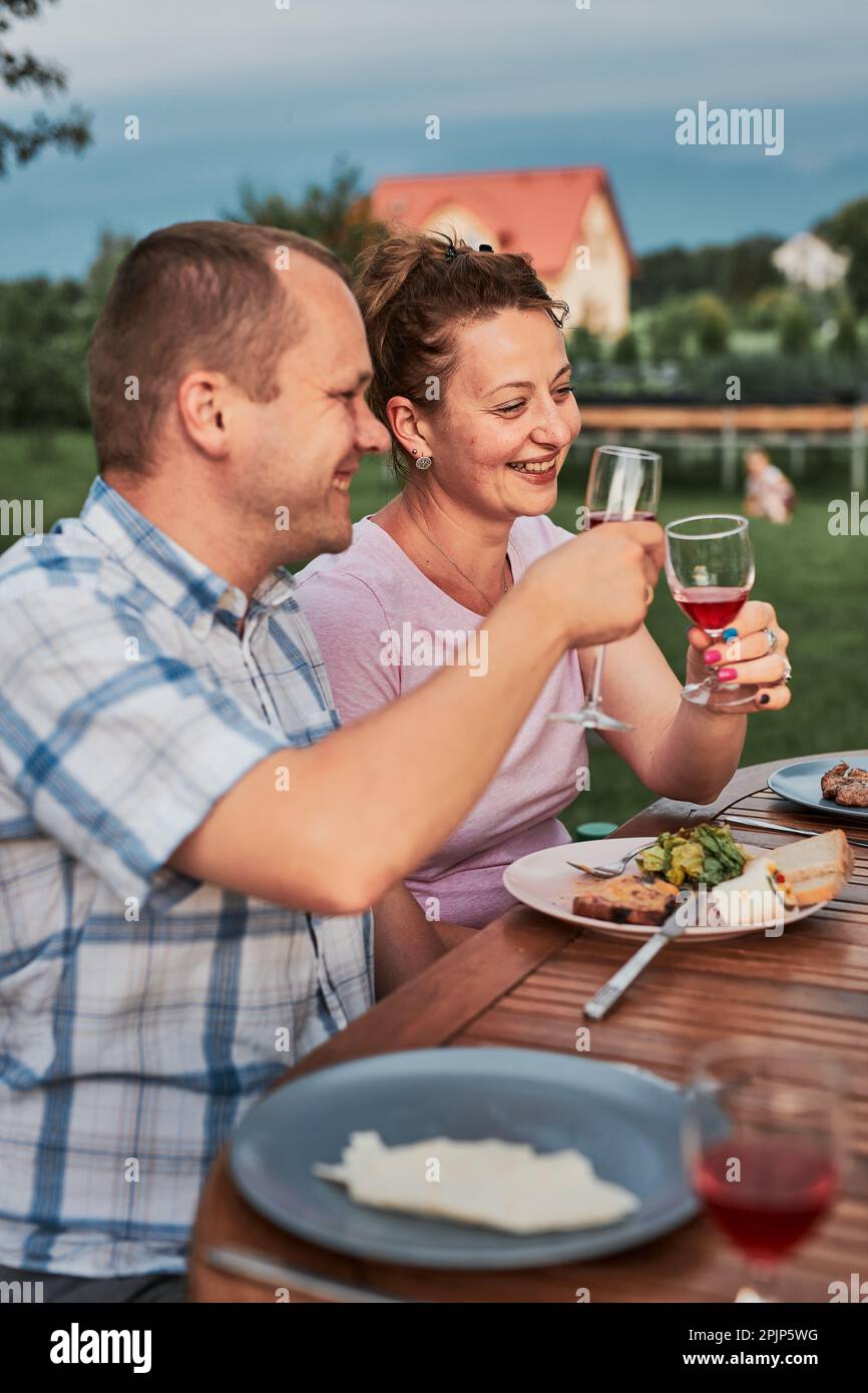 Familia haciendo brindis durante el picnic de verano cena al aire libre en un jardín casero. Cerca de la gente que sostiene copas de vino con vino tinto Foto de stock
