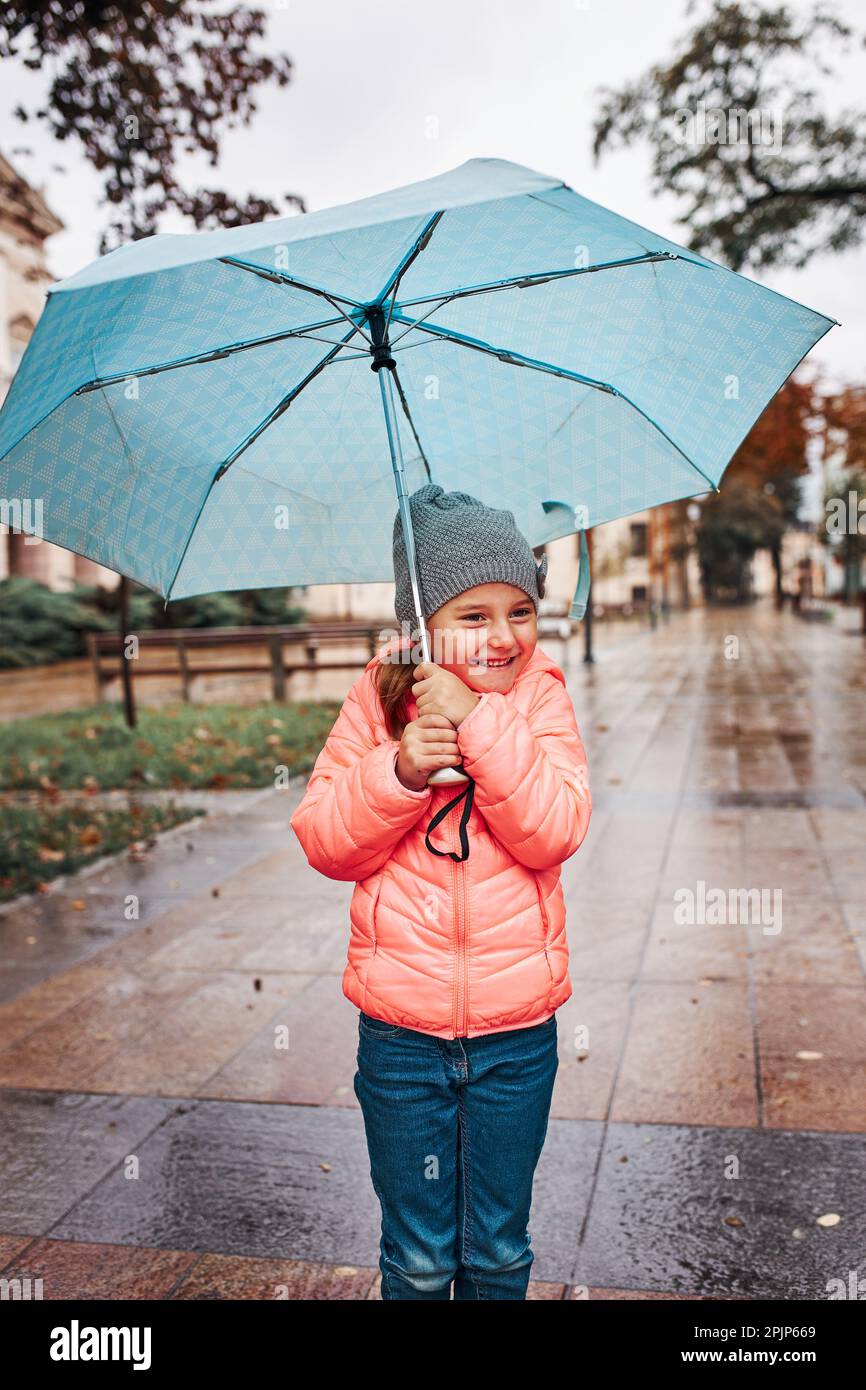 Pequeña chica sonriente feliz sosteniendo un paraguas azul grande durante la caminata en un día de otoño sombrío y lluvioso Foto de stock