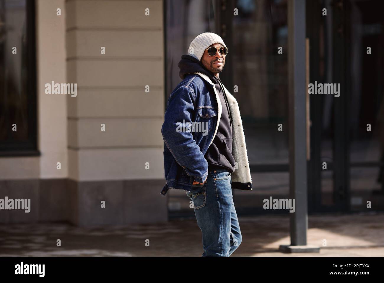 Guapo modelo de hombre afroamericano con elegante chaqueta vaquera, sombrero, sudadera con capucha y gafas sol caminando en la calle de ciudad Fotografía de stock