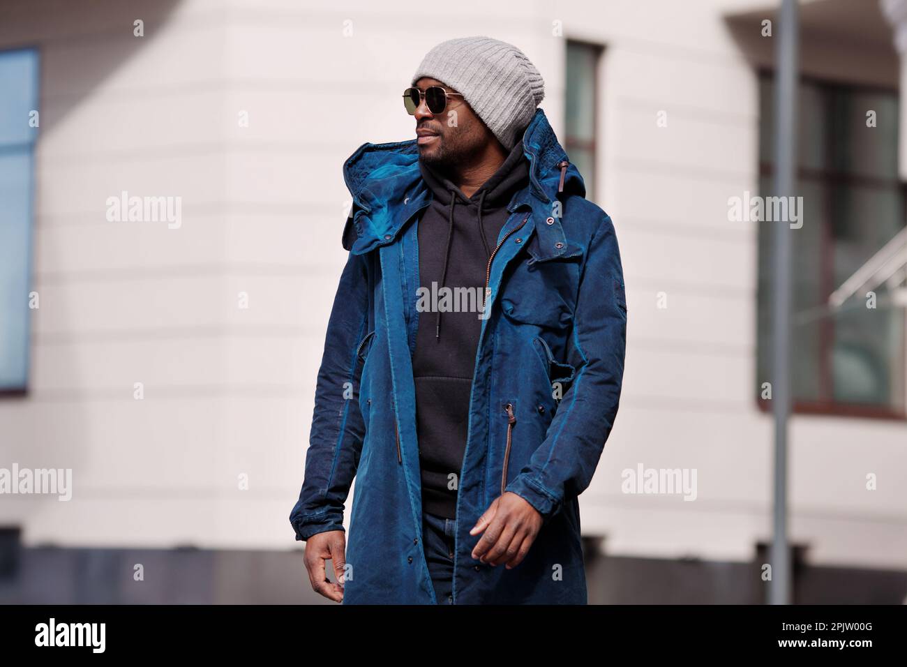Retrato auténtico del hombre afroamericano guapo que camina en la ciudad,  vistiendo elegante abrigo parka del traje, sudadera con capucha, sombrero  de punto y gafas de sol. street sty Fotografía de stock 